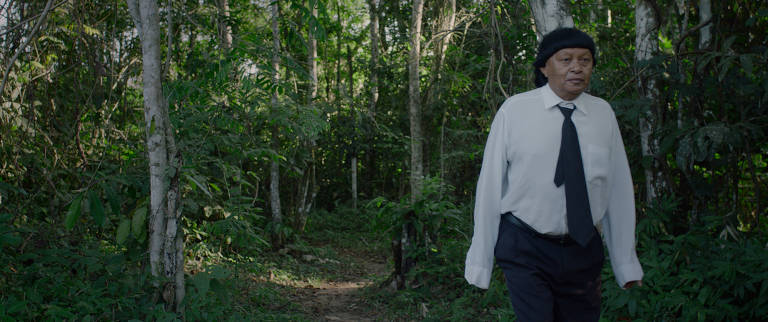 Cena do filme 'Ex-Pajé'; seu protagonista, Perpera Suruí, ex-pajé de sua aldeia, está com coronavírus; vemos um homem com o cabelo cortado à moda indígena, porém com camisa branca, calça e gravata pretas, andando na floresta