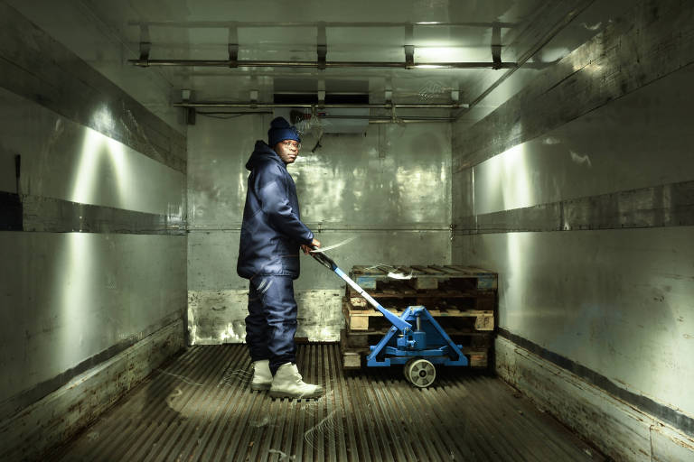 Retrato do camaronês Kouotou Yamaya Mouhamed, que trabalha em caminhão frigorífico de empresa logística, que usa agasalho e botas brancas, além de óculos de proteção. Opera uma máquina dentro de um caminhão.