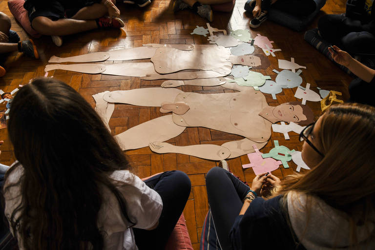Crianças no chão em volta de recortes de papel que mostram como é o corpo humano