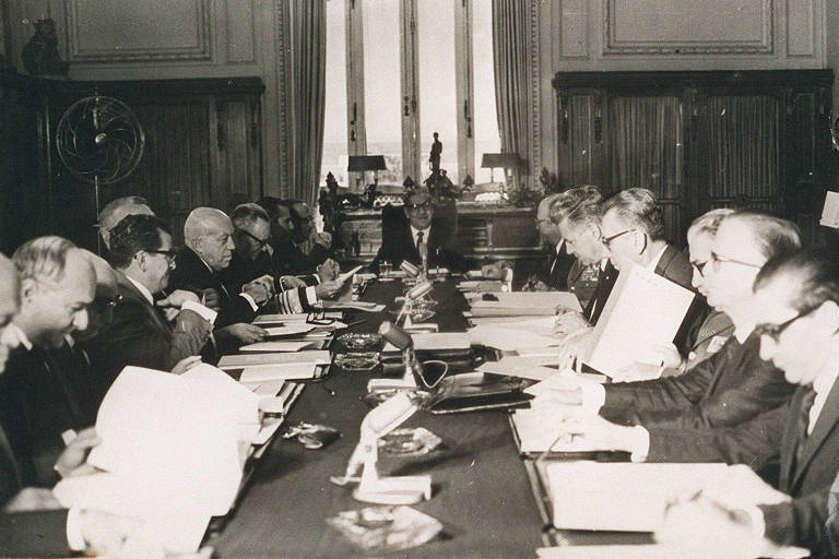 foto em preto e branco mostra vários homens brancos, vestidos com terno ou farda, sentados ao redor de uma mesa comprida de madeira 