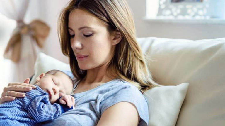 Mulher loira segura um bebê recém-nascido em seu peito. Ela veste azul e o bebê também