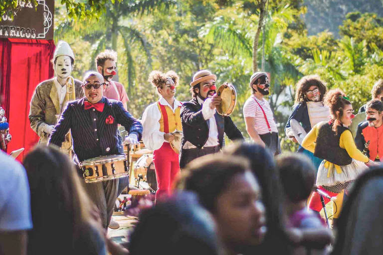 Artistas do Circo Teatro Palombar se apresentam com os rostos pintados, roupas de palhaço e tocando instrumentos enquanto o público os assiste