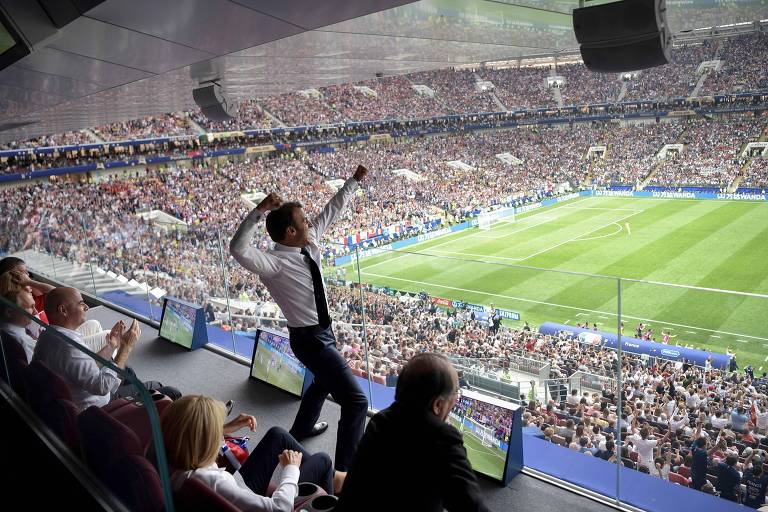 Macron, de camisa branca e gravata escura, ergue os braços e vibra em espaço reservado no estádio Luzhniki, que está lotado durante a final da Copa de 2018