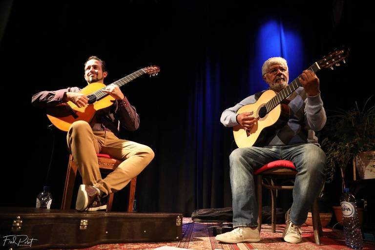 Violonistas Dinho Nogueira (à esquerda) e Zé Barbeiro (à direita) se apresentam sentados, com seus violões, em show