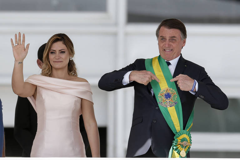 Michelle, mulher branca de cabelos loiros de lado, usa vestido claro e acena com a mão direita; à esq. dela, Bolsonaro apontando com as duas mãos para a faixa presidencial, em seu peito