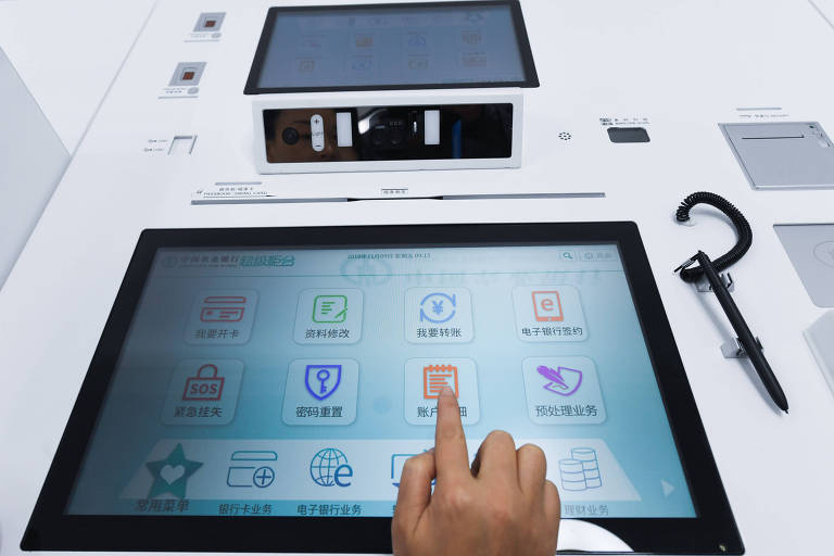 Na foto, uma máquina inteligente de autoserviço bancário aparece com a tela disposta. Vários ícones com escritas em chinês estão dispostos pela tela enquanto a mão de uma pessoa aparece clicando em um deles.