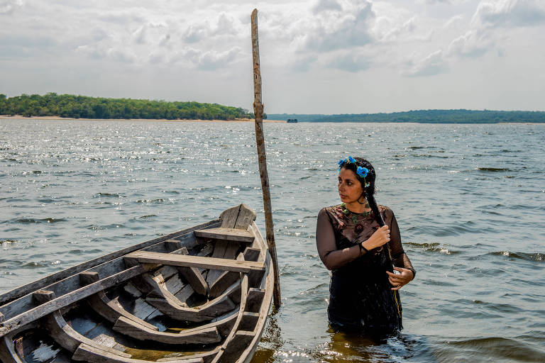 A pianista Carla Ruaro, vestida de preto, mexe em sua trança; vestida de preto, ela está ao lado de um barco simples e com água até a cintura no rio Arapiuns, no Pará