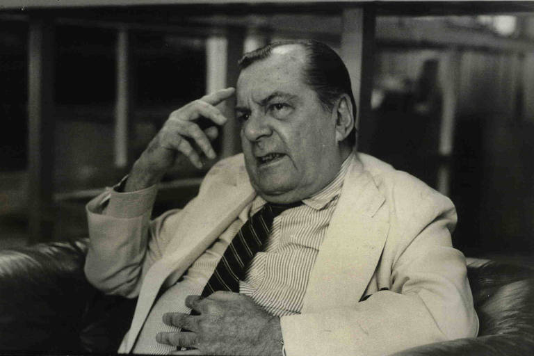 O jornalista Joel Silveira, sentado de lado, com uma das mãos na cabeça,, durante entrevista em 1985