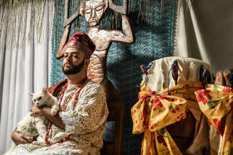 babalorixá Diego Montone, 33, em suas vestes religiosas, sentado com gato no colo, e ao lado de um tambor 