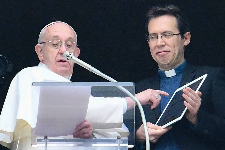 O papa, vestido de branco e em frente à uma tribuna com um microfone em frente, aponta para um tablet segurado por um padre, enquanto apresenta o app  Click to Pray, ou Clique para Rezar 