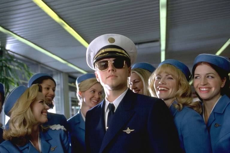 um homem caracterizado como piloto de avião está cercado de atendentes de bordo na cena de um filme