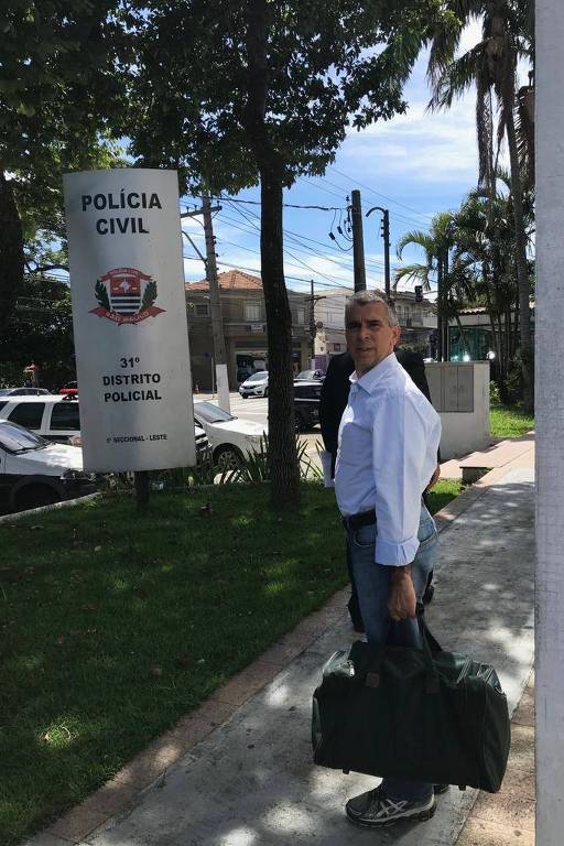 De camisa clara, Ronilson Rodrigues posa ao lado da placa de entrada de delegacia em São Paulo, com uma sacola de viagem na mçao