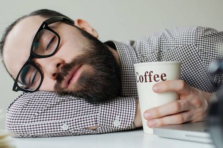 Homem branco, de óculos e barba, dorme debruçado sobre uma mesa; ele segura um copo de café em uma das mãos