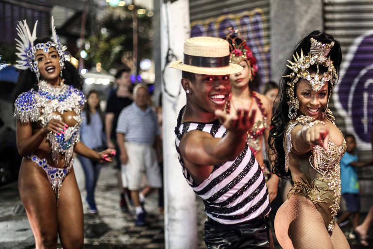 Sambista de chapéu de palhinha estende a mão para a frente ao lado de moça com fantasia de escola de samba