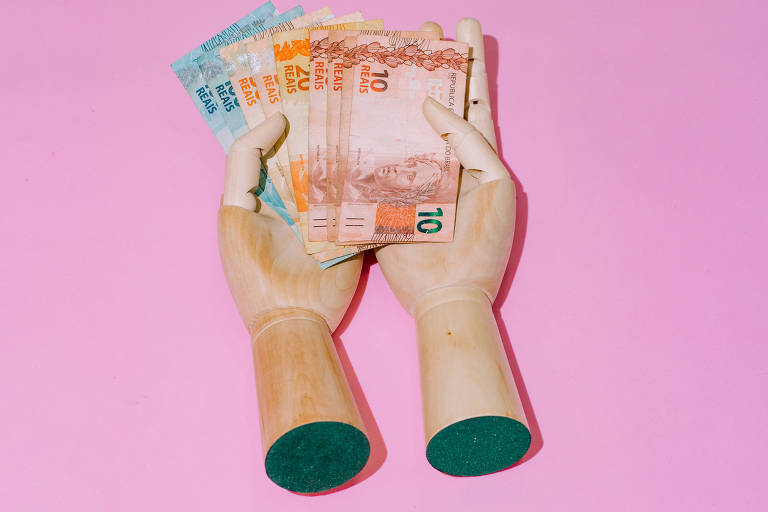 Duas mãos de madeira sobre fundo rosa seguram notas de R$ 100, R$ 50 e R$ 10