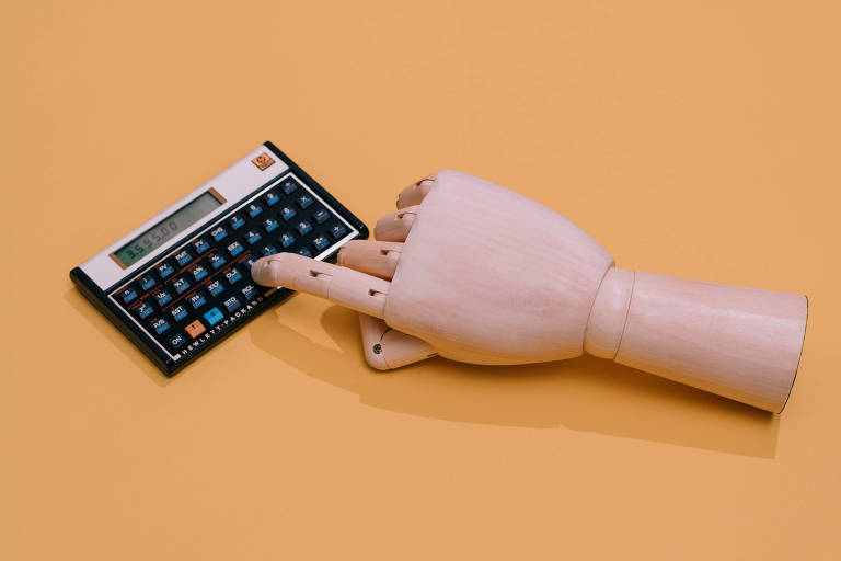 Mão de madeira mexe em calculadora científica. Imagem sobre fundo laranja.