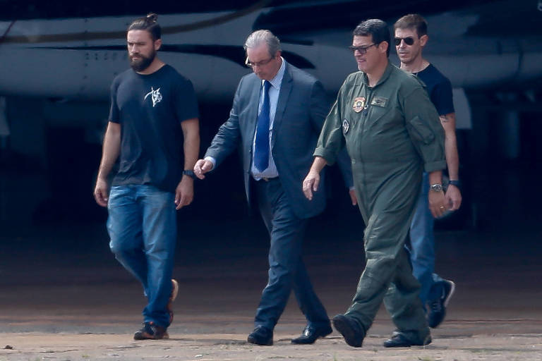 deputado eduardo cunha de terno e gravata é escoltado por três policiais, de camisa preta; dois pilotos com macacão verde estão na cena, com avião atrás