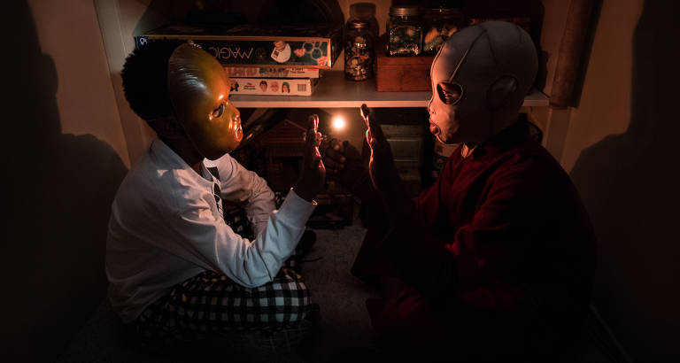 Dois meninos negros, mascarados, olhando um para o outro, enquanto carregam um palito com uma luz