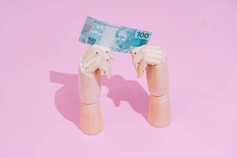 Foto mostra um fundo rosa com duas mãos articuladas de madeira segurando uma nota de cem reais aberta