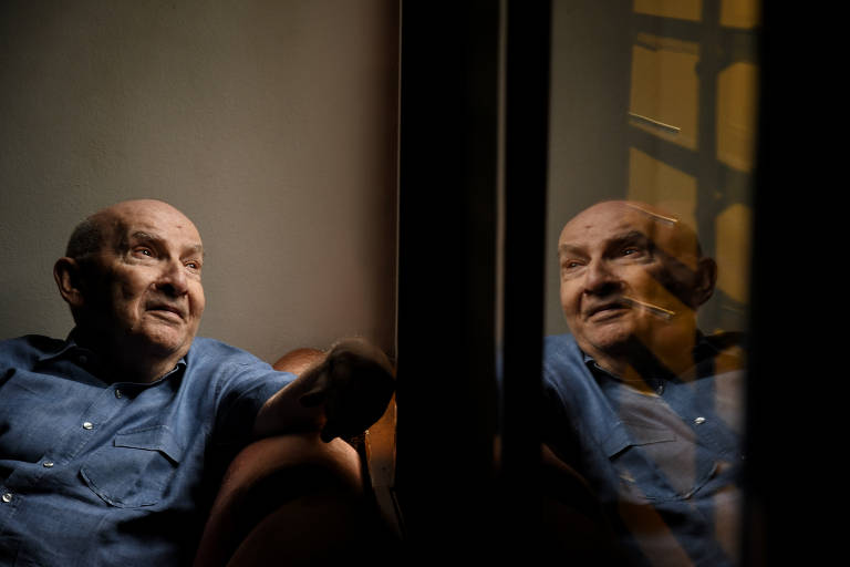 foto colorida de homem branco careca, idoso, veste camisa azul e está sentado em uma poltrona. ao lado dele, um vidro reflete sua imagem 