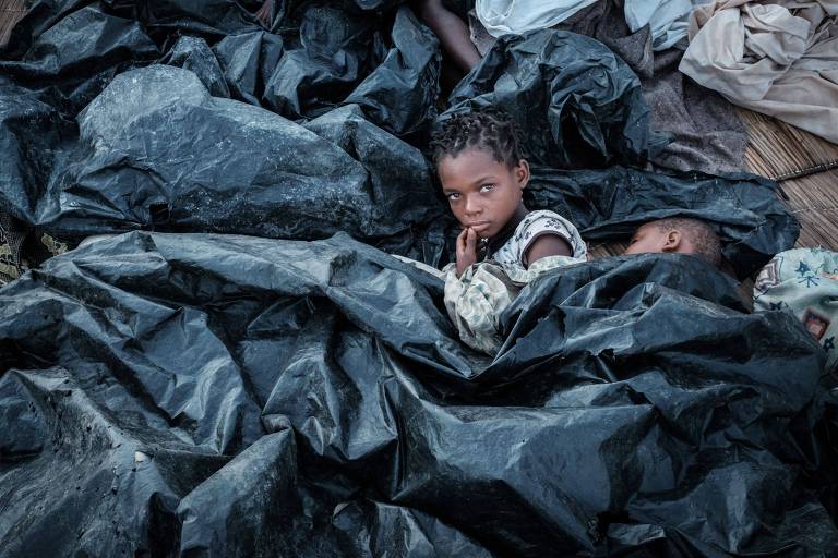 Uma menina coberta por plástico preto olha em direção à câmera, enquanto ao seu lado uma criança menor dorme