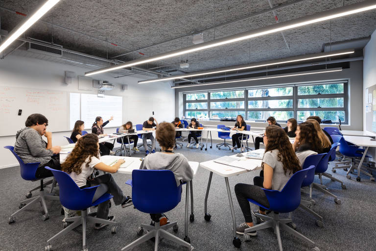 estudantes estão sentados em mesas de duas pessoas, dispostas em círculo numa sala de aula; atrás da professora, um texto é projetado numa tela