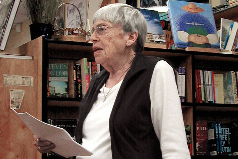 A escritora Ursula K. Le Guin lê páginas em sua mão e apoia a outra mão sobre uma mesa