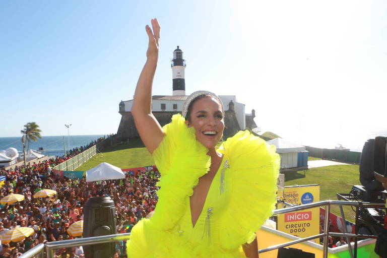 Ivete Sangalo no trio em Salvador no Carnaval 2019. A cantora está com um braço levantado, com uma fantasia amarela, sorrindo, com o Farol da Barra ao fundo. 