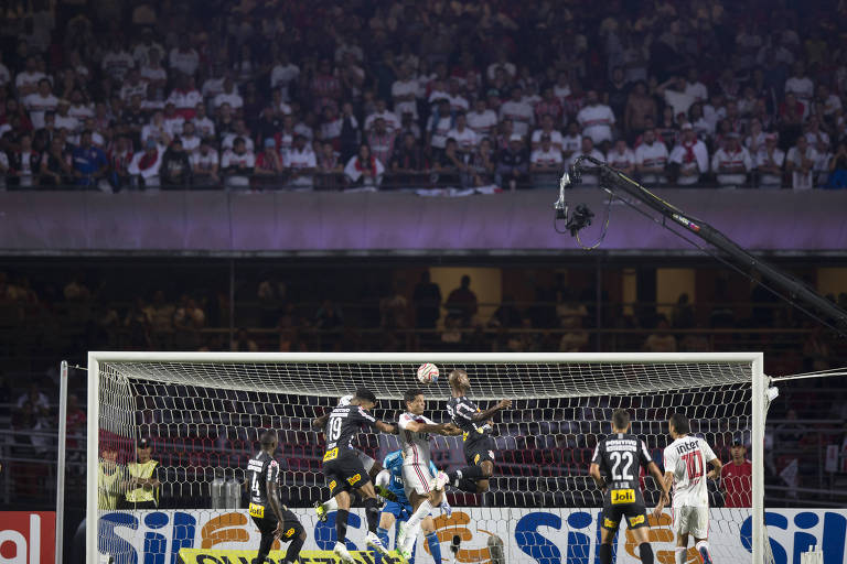 Homens com roupas brancas e pretas cabeceiam bola em direção a gol, em estádio 