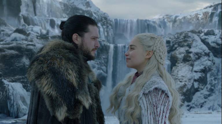 A imagem mostra os personagens Jon Snow e Daenerys Targaryen, da série  'Game of Thrones', em um cenário de inverno, com uma cachoeira ao fundo. Eles estão se encarando, ambos vestindo roupas pesadas e com detalhes de pele animal. O ambiente é coberto de neve e gelo, criando uma atmosfera fria e dramática