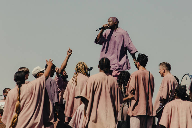Kanye com um túnica roxa em um palco com pessoas com túnicas de um tom mais claro em volta dele