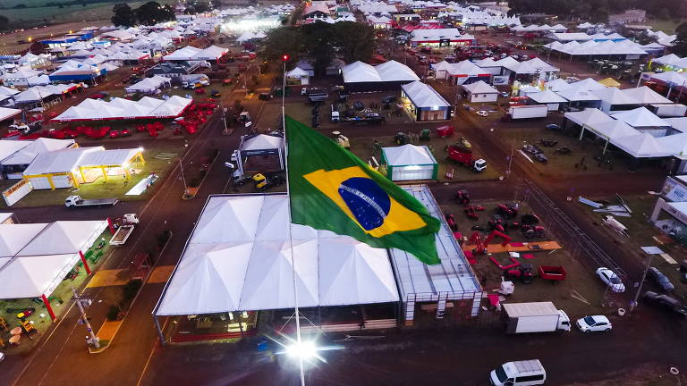 Imagem mostra uma bandeira do Brasil em primeiro plano, com uma série de estandes ao fundo, que abrigam expositores da Agrishow, em Ribeirão Preto