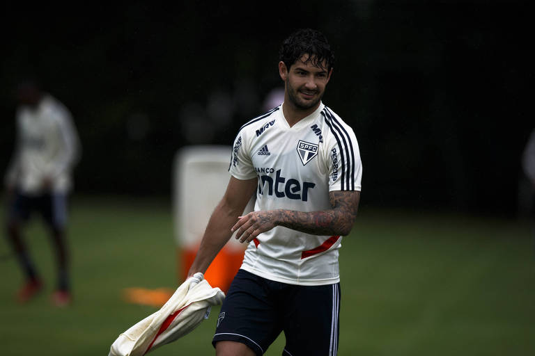 Carregando uma blusa branca com a mão direita, o sorridente atacante Alexandre Pato caminha em um dos gramados do centro de treinamento do São Paulo