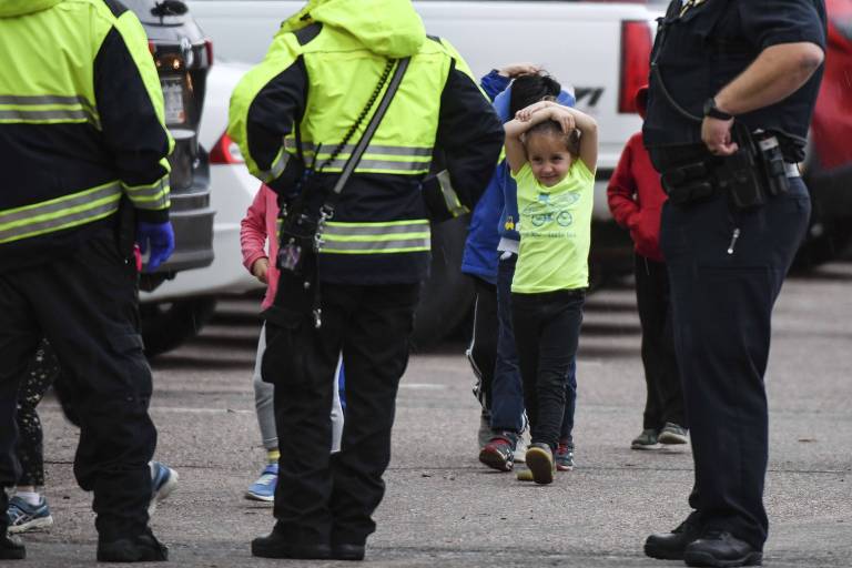 crianças andam em fila com as mãos na cabeça; policiais observam