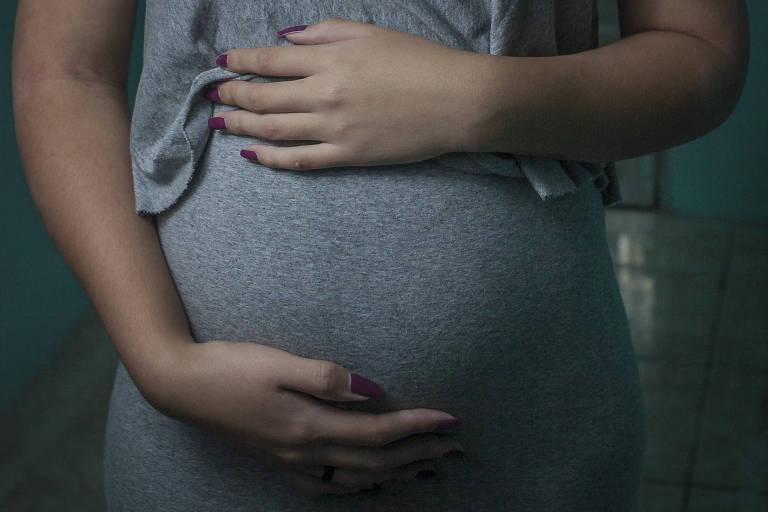 Imagem foca a barriga de uma mulher grávida; ela está de vestido cinza e segura a barriga com as mãos