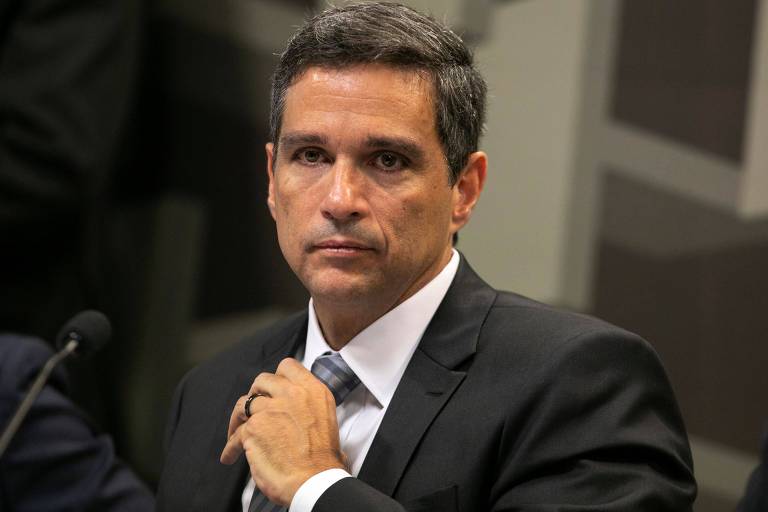 O presidente do Banco Central, Roberto Campos Neto, aparece em frente a um pequeno microfone. Na foto, ele olha sério para a câmera enquanto ajeita casualmente a gravata que usa.
