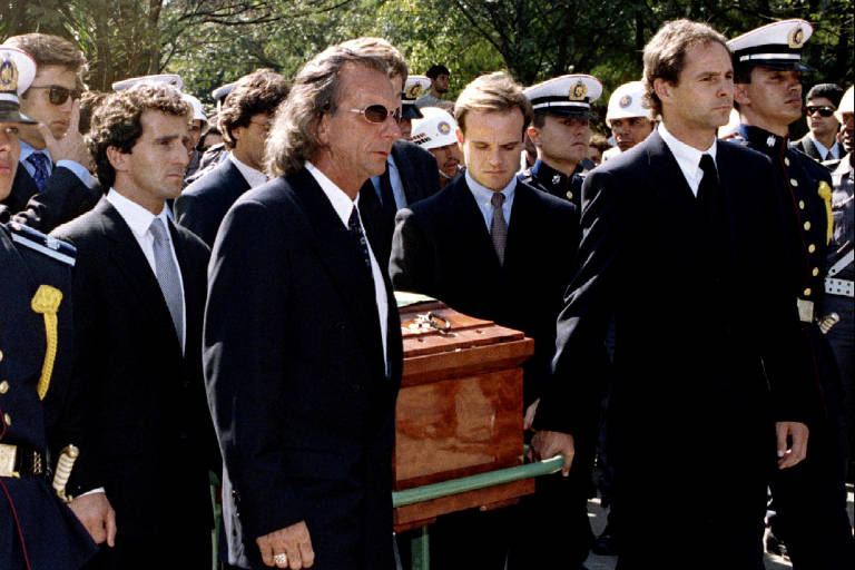 A imagem mostra um grupo de homens vestidos de terno preto， carregando um caixão de madeira. Eles estão cercados por uma multidão， incluindo membros de forças armadas， que observam a cerimônia. O ambiente parece ser ao ar livre， com árvores ao fundo.