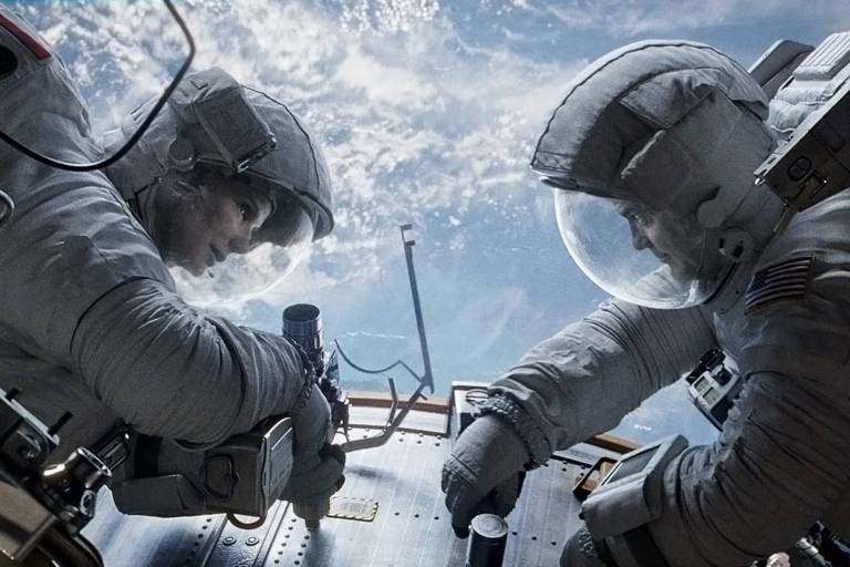 Sandra Bullock, à esquerda, e George Clooney, à direita estão flutuando no espaço no papel de seus personagens em "Gravidade". Eles se encaram e vestem roupas brancas de astronauta. Ao fundo da imagem é possível ver uma parte do planeta Terra. 