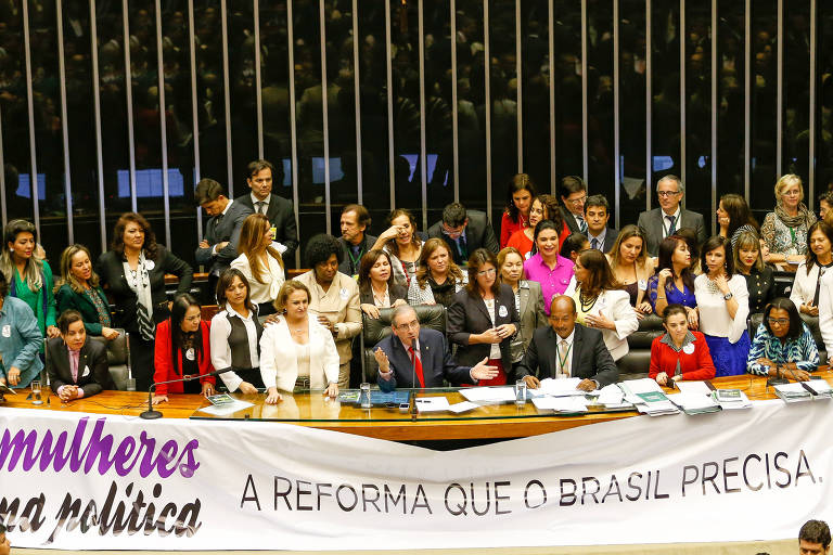 Uma mesa comprida de madeira, com uma multidão de mulheres aglomeradas atrás de um homem branco de cabelos brancos e óculos. Na frente, uma faixa onde se lê: "mulheres na política, a reforma que o Brasil precisa"