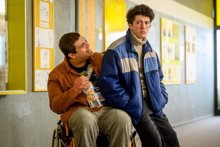 Garoto sentado na cadeira de rodas e amigo dele sentado na  ponta da cadeira de rodas, em cena do filme "Como Vender Drogas Rápido"