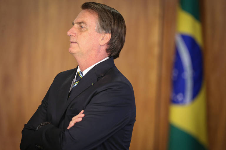 Jair Bolsonaro de perfil e braços cruzados. Uma bandeira do Brasil aparece ao fundo