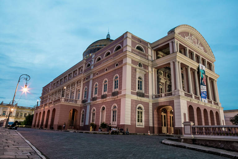 Imagem do Teatro Amazonas， um edifício histórico localizado em Manaus， Brasil. A foto mostra a fachada do teatro， que apresenta uma arquitetura imponente com detalhes em cores claras e escuras. O céu está nublado， e há iluminação pública visível ao redor do edifício.