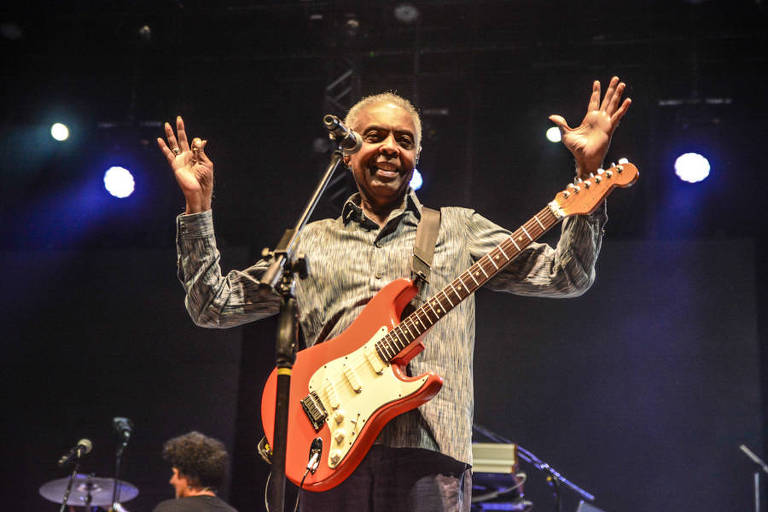 Cantor Gilberto Gil se apresenta em show. Na imagem, ele está com os dois braços levantados, diante de um microfone, enquanto olha para baixo e sorri. Segura uma guitarra vermelha e branca e veste camisa social cinza, de manga longa