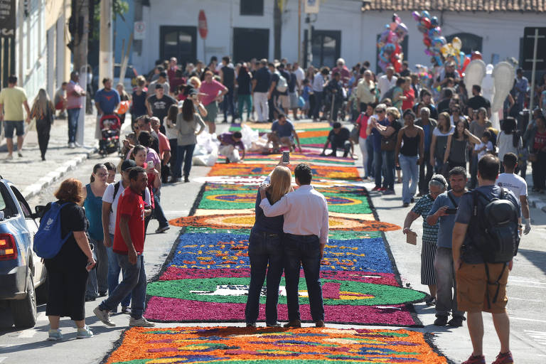 Pessoas andam em procissão na cidade de Santana do Parnaíba nas celebrações de Corpus Christi. Ao centro da foto, um homem e uma mulher de costas se abraçam. Eles estão sobre um tapete colorido com motivos religiosos que enfeita a rua.