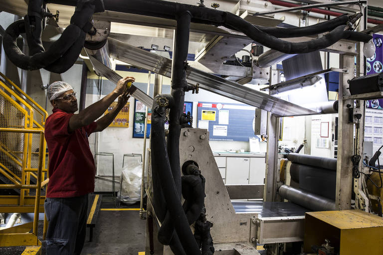 Um operário está ajustando uma máquina em uma fábrica. Ele usa uma camisa vermelha, óculos de proteção e um touca. A máquina é composta por várias partes metálicas e tubos, com rolos de material em funcionamento. Ao fundo, há uma escada amarela e uma parede com documentos e gráficos visíveis.