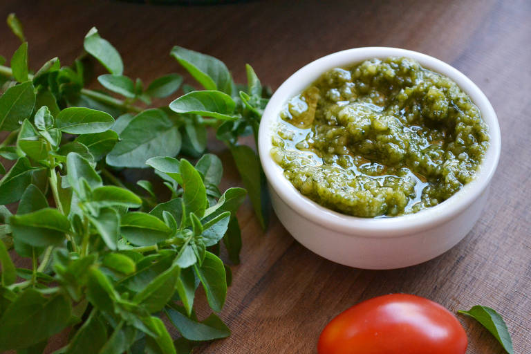 A imagem mostra um pequeno recipiente branco com molho pesto， ao lado de ramos de orégano fresco e um tomate vermelho. O fundo é uma superfície de madeira.