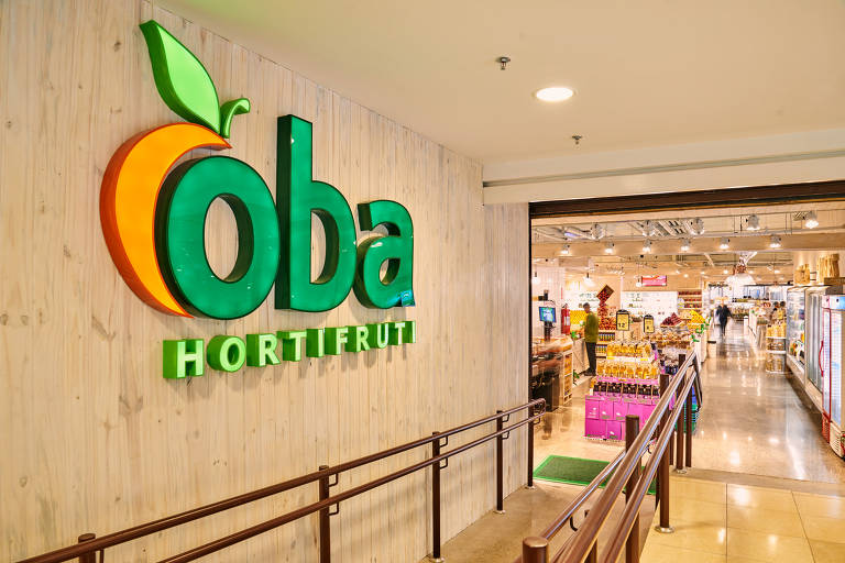 A imagem mostra a entrada de um supermercado chamado Oba Hortifruti. O logotipo é destacado em cores vibrantes， com a palavra 039;Oba039; em verde e laranja， acompanhada de uma folha verde. Ao fundo， é possível ver o interior do supermercado， com prateleiras e produtos expostos.
