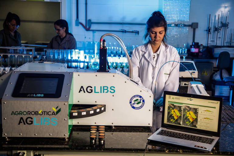 Uma cientista, de jaleco, de pé próxima a um computador e uma máquina em um laboratório 