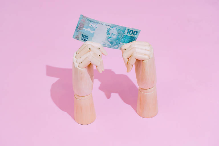 Uma foto com o fundo rosa mostra duas mãos articuladas de madeira segurando uma nota de 100 reais.