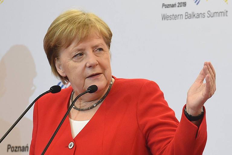 A chanceler alemã, Angela Merkel, aparece sentada frente a uma mesa com microfones. Ela veste um blazer vermelho e olha para a esquerda, fazendo um gesto de aproximação